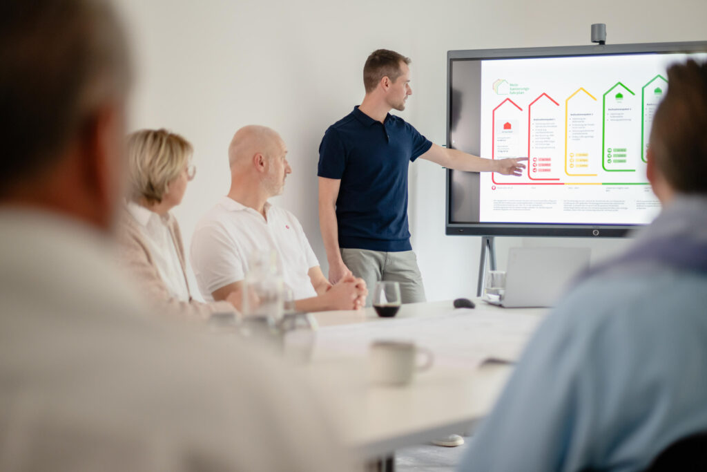 Energieberater steht vor Smartboard und erklärt den Sanierungsfahrplan. Im Vordergrund sieht man Menschen an einem Tisch sitzen.